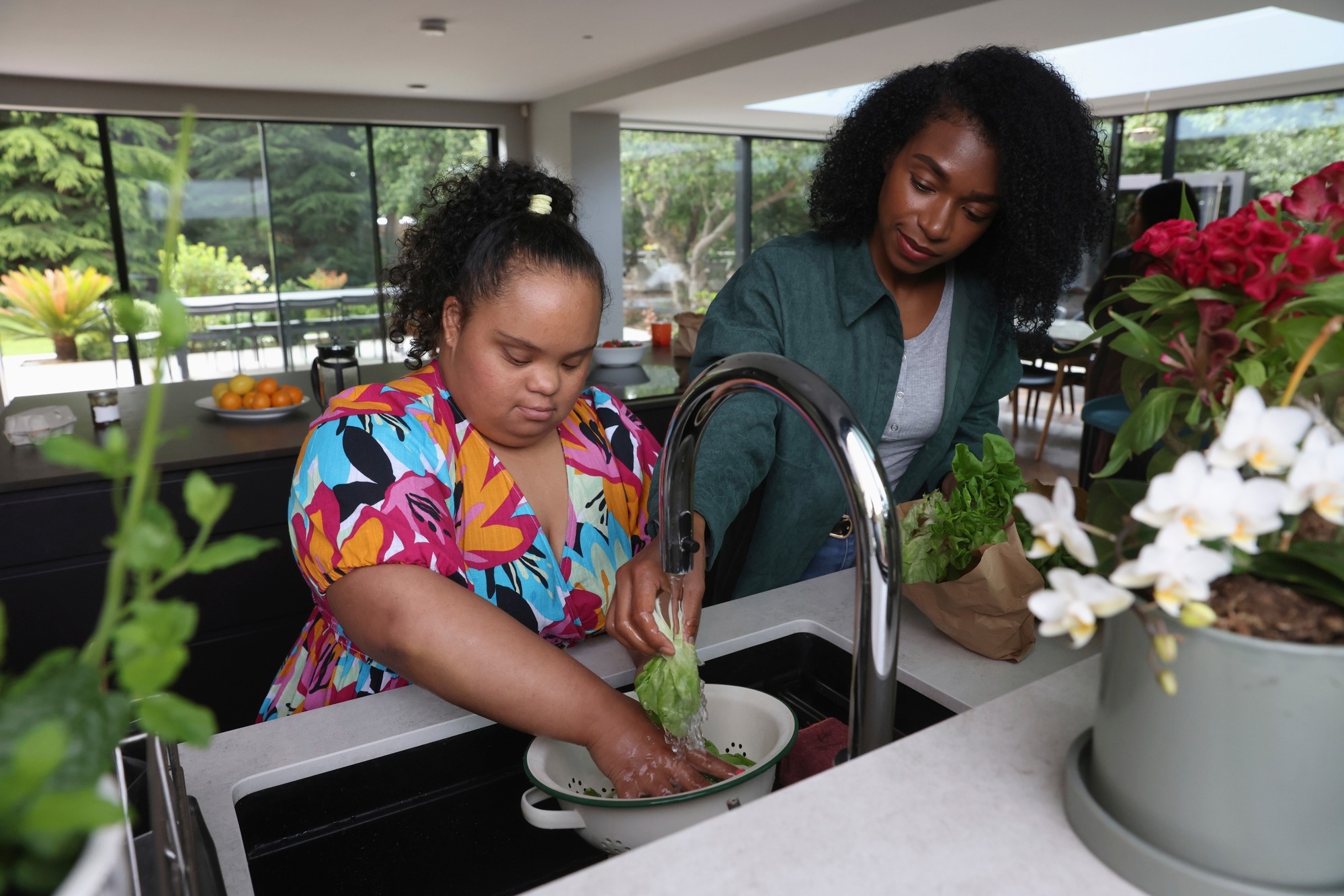 Two women wash lettuce together in a bright kitchen, emphasizing teamwork and healthy cooking. Fresh vegetables and plants are visible, highlighting a vibrant home environment.