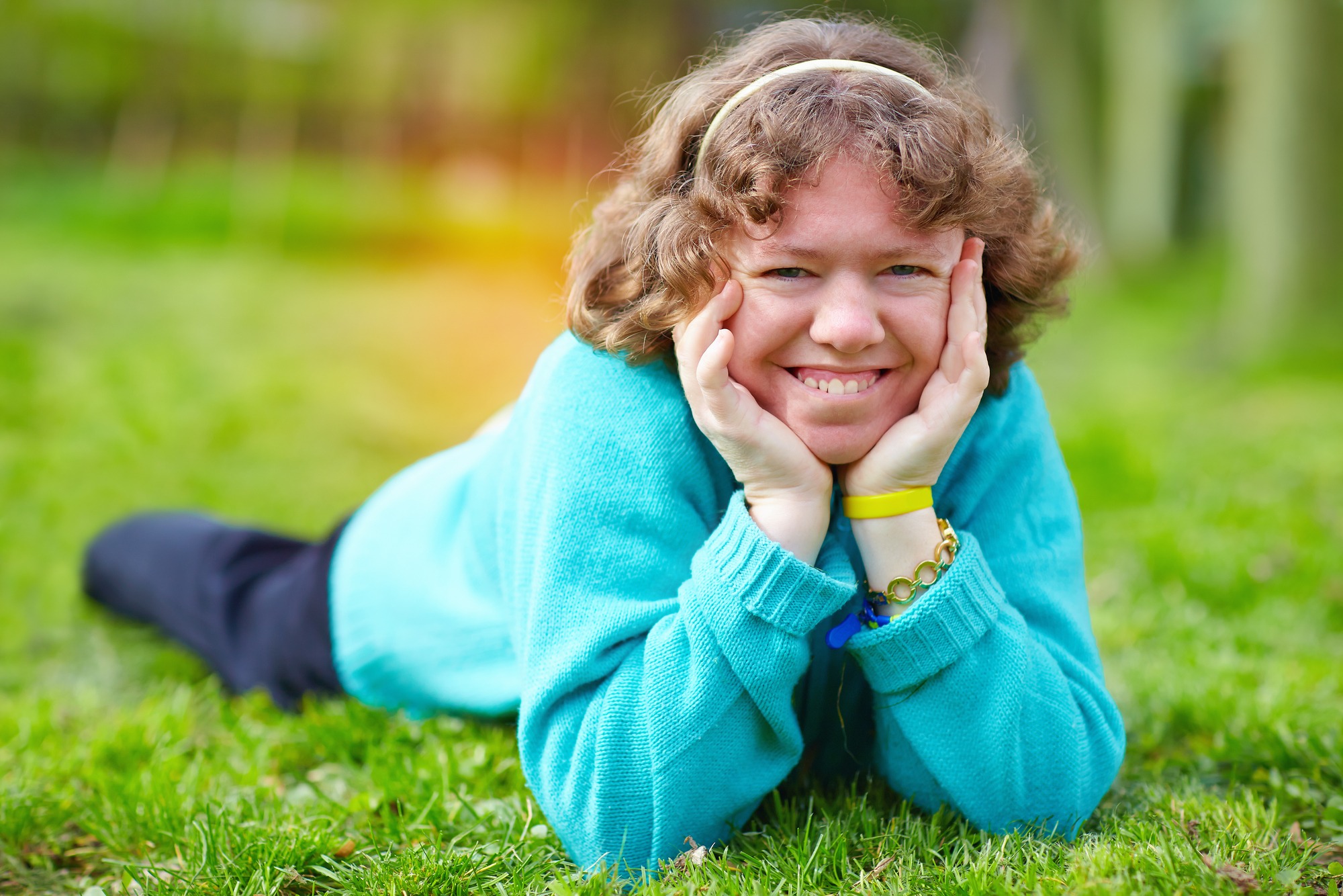 A young woman with curly hair smiles while resting on her stomach in a grassy area, wearing a bright blue sweater and bracelets.